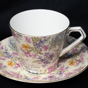 Op de afbeelding: Een theekopje en schotel met bloemenmotief met een delicaat bloemenpatroon in roze, paarse en gele tinten op een crèmekleurige achtergrond. Het kopje en de schotel hebben een gouden rand.