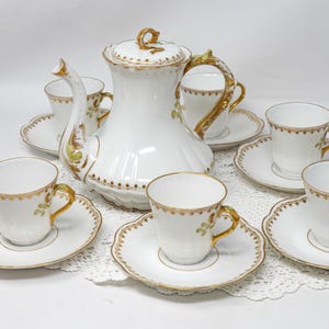 May include: A white porcelain tea set with gold trim. The set includes a teapot with a lid, six teacups, and six saucers. The cups and saucers have a scalloped edge. The teapot and cups have gold handles.