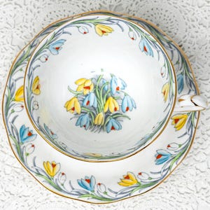 May include: A white porcelain teacup and saucer set featuring a delicate hand-painted floral pattern of yellow and light blue crocus flowers with green stems. The rim of the cup and the edge of the saucer are accented with a thin gold trim.