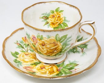 May include: A vintage teacup and saucer set. The white porcelain cup and saucer feature a yellow rose floral design with green leaves. The edges are trimmed with gold. A classic tea set for serving tea.