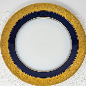 May include: A round, white plate with a navy blue band and a gold rim. The gold rim features a detailed, repeating leaf pattern. The plate is set on a white, lacy surface.