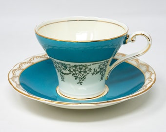 May include: A teal teacup and saucer set with a gold trim and a white floral design. The cup has a wide, flared rim and a delicate handle.
