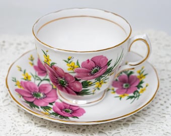 May include: A white teacup and saucer set with a floral pattern. The cup and saucer feature pink and yellow flowers with green leaves on a white background. The edges of the cup and saucer are trimmed with gold.