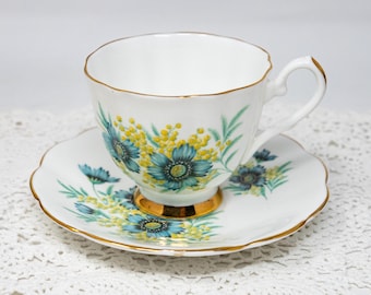May include: A white teacup and saucer with a floral pattern. The pattern features blue and yellow flowers with green leaves. The cup and saucer have a gold trim.