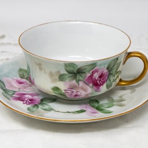 May include: A white porcelain teacup and saucer with a gold rim and floral design. The cup has a gold handle and the saucer features pink roses and green leaves.