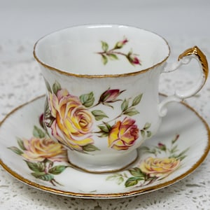 Peut inclure: Une tasse à thé et une soucoupe blanches avec une bordure dorée et un motif floral représentant des roses jaunes et roses.
