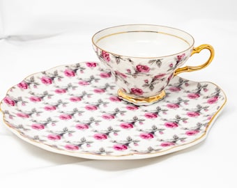 Pode incluir: Um conjunto de chávena e pires de chá branco com um padrão de rosas cor-de-rosa e guarnição dourada. A chávena e o pires são ambos decorados com um padrão repetido de rosas cor-de-rosa e folhas verdes.
