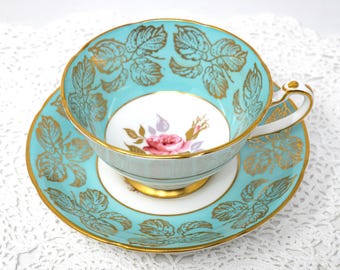 May include: A vintage teacup and saucer set. The teacup and saucer are a vibrant turquoise color with gold leaf detailing. The teacup interior features a floral design with a pink rose. The set is trimmed with gold accents, creating an elegant and classic look.