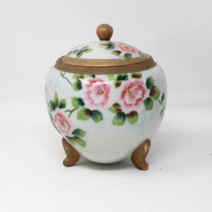 May include: A white porcelain jar with a lid and three feet. The jar is decorated with pink and green floral designs.
