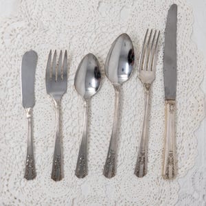 May include: A set of five silver-plated cutlery pieces, including a butter knife, fork, and three spoons, all with ornate designs. The cutlery is arranged on a white lace tablecloth.