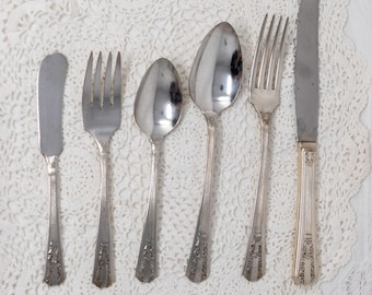 Può includere: Un set di cinque posate argentate, tra cui un coltello da burro, una forchetta e tre cucchiai, tutti con disegni decorativi. Le posate sono disposte su una tovaglia di pizzo bianco.