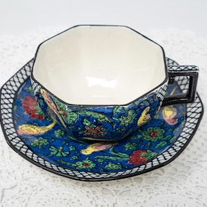 May include: A vintage teacup and saucer set. The cup and saucer feature a dark blue background with a floral and bird design in red, yellow, and green. The cup has a geometric shape and a black handle. The saucer has a black and white border.