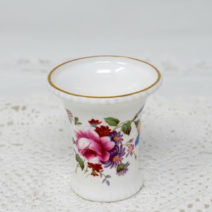 May include: A small white porcelain vase with a gold rim and a floral pattern. The vase features a pink rose, purple and white flowers, and green leaves.