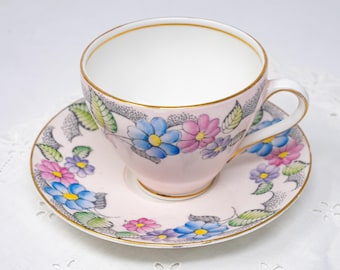 May include: A pink teacup and saucer with a floral pattern. The cup and saucer are decorated with a delicate design of pink, blue, and yellow flowers with green leaves. The rim of the cup and saucer is trimmed with gold.