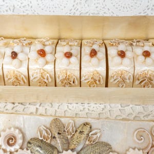 May include: A rectangular wooden box containing eight rectangular bars of cream-colored soap. Each soap bar is decorated with a floral design made of small white shells and a single amber-colored bead in the center, with additional shell accents.