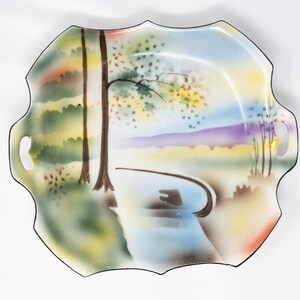 May include: A white ceramic serving dish with a scalloped edge and two handles. The dish features a hand-painted scene of a forest with trees, a river, and a bridge. The colors are muted and include shades of green, blue, yellow, and purple.