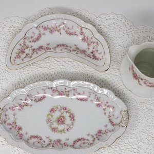 Puede incluir: Un juego de té de porcelana vintage con una fuente, un plato pequeño y una jarra para la crema. Las piezas blancas están decoradas con motivos florales rosas y ribetes dorados. El juego se exhibe sobre un tapete de encaje blanco.
