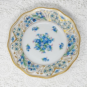 May include: A white porcelain plate with a gold rim and a delicate floral design. The plate features a central bouquet of blue flowers and a border with intricate cut-out patterns and additional blue floral accents. The plate is approximately 20 cm in diameter.