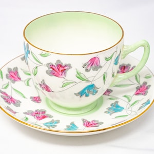 Puede incluir: Una taza de té y un platillo blancos con un patrón floral en tonos de rosa, azul y gris. La taza tiene un interior verde claro y un borde dorado.
