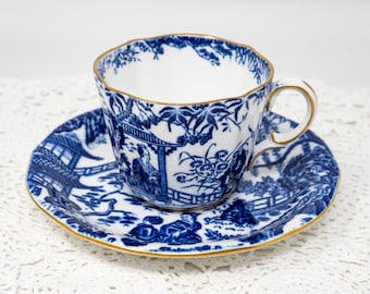 May include: A blue and white porcelain teacup and saucer set. The cup and saucer feature a detailed landscape design with gold trim. The teacup has a scalloped rim and a curved handle. The set is placed on a white surface.