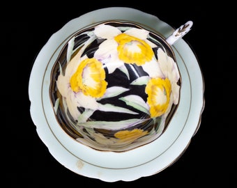 May include: A vintage teacup and saucer set with a black background and a floral design of yellow daffodils. The cup and saucer are trimmed with gold.