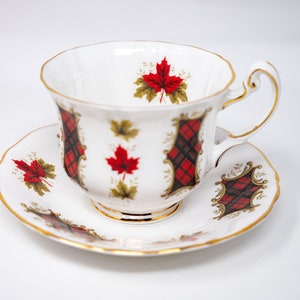 May include: A white teacup and saucer set with a red and black plaid and maple leaf design. The set has a gold trim.