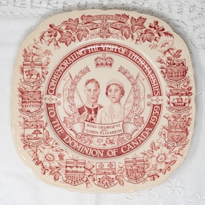 May include: A white ceramic plate with a red and white design commemorating the visit of King George VI and Queen Elizabeth to Canada in 1939. The plate features the coats of arms of the Canadian provinces.