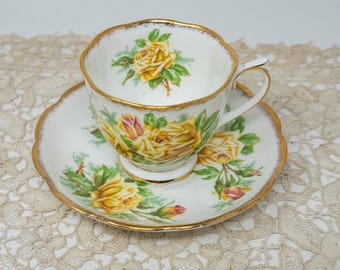 May include: A white teacup and saucer set with a gold rim and yellow rose floral design. The teacup has a curved handle and sits on a matching saucer. The set is on a lace tablecloth.
