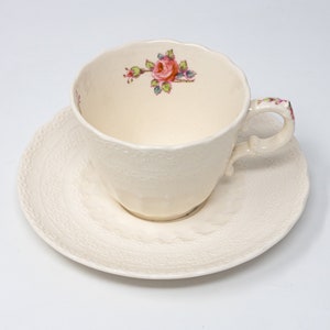 May include: Cream-colored teacup and saucer set. The teacup features a floral design with pink and red roses. The saucer and cup have a textured, dotted pattern. The handle of the cup is also decorated with floral accents.