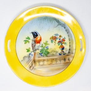 May include: A yellow ceramic serving plate with two handles features a painted design of a blue and red bird perched on a branch with a vase of flowers in the background.
