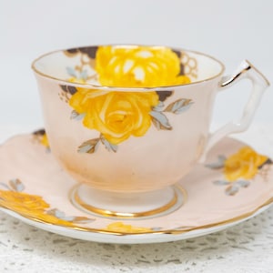 May include: A pink teacup and saucer set featuring a yellow rose floral design. The teacup has a dainty handle and the saucer has a scalloped rim.