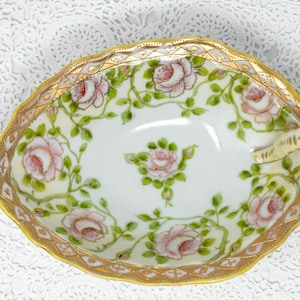 May include: Oval-shaped, antique porcelain dish with a gold rim and handle. The dish is decorated with pink roses and green leaves on a white background. The rim has a gold trim with a scalloped edge. A decorative piece for serving or display.