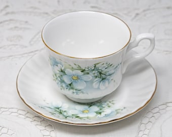 May include: A white teacup and saucer with a delicate floral pattern featuring blue flowers and green leaves. The cup and saucer have a gold trim.