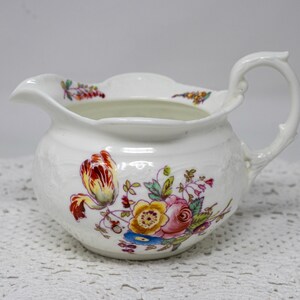 May include: White ceramic creamer with a floral design. The creamer features a curved handle and a spout for pouring. The floral pattern includes red, yellow, pink, and blue flowers with green leaves. The creamer has a textured surface.