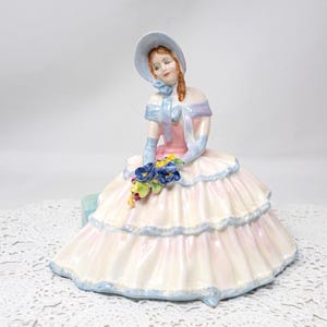 May include: A porcelain figurine of a woman in a layered pink and white dress, holding a bouquet of blue and yellow flowers. She wears a light blue hat, gloves, and accents on her dress. A vintage collectible figurine.
