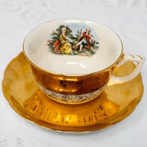 May include: A gold-rimmed teacup and saucer set with a painted scene of two people in a garden. The saucer has a gold rim and a gold-colored interior.