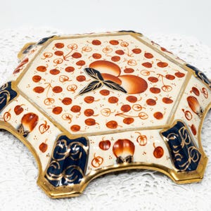 May include: An octagonal ceramic dish with a cream background, decorated with orange and red fruit motifs. The dish has gold trim and navy blue accents. The design includes a cluster of orange fruits and a black leaf.