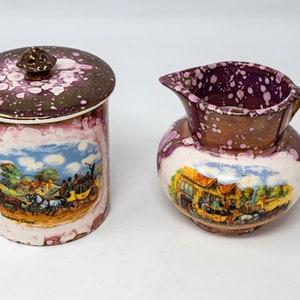 May include: A vintage cream and purple ceramic creamer and sugar bowl set with a gold trim. The creamer has a handle and the sugar bowl has a lid. Both pieces feature a painted scene of a horse-drawn carriage and a building with the text "Docken's Dove".