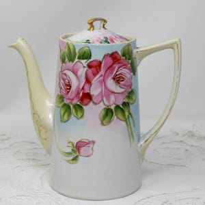 Puede incluir: Una cafetera de porcelana blanca con ribete dorado y un diseño floral con rosas rosas y hojas verdes. La cafetera tiene una tapa con un pomo dorado.