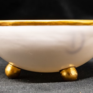 May include: A white ceramic bowl with a gold rim and three gold feet.