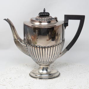 May include: Silver teapot with a black handle and a fluted design. The teapot has a round body and a tall, slender spout.