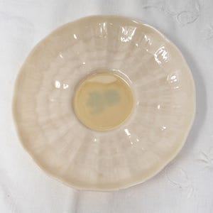 May include: A white ceramic saucer with a scalloped edge and a slightly raised center. The saucer has a subtle, textured pattern.