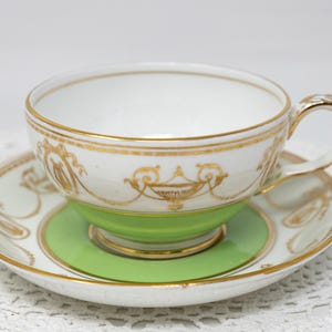 Könnte beinhalten: Weißes Tee-Set mit goldenen Verzierungen. Die Tasse hat einen goldenen Henkel und einen grünen Sockel. Die Untertasse ist weiß mit goldenem Rand und einem grünen Mittelteil. Das Set ist wahrscheinlich Vintage oder antik.