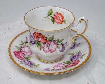May include: A white teacup and saucer set with a floral design. The cup and saucer are adorned with pink, purple, and red flowers with green leaves. The rims are trimmed with gold. The set is on a white surface.