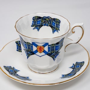 Könnte beinhalten: Ein weißes Tee-Set mit einem blauen und grünen Tartanmuster und goldenen Verzierungen. Die Tasse zeigt ein schottisches Wappen mit einem Löwen und einem Schild.
