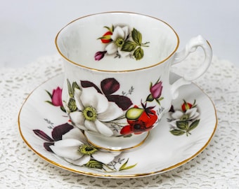 May include: A white teacup and saucer with a floral design. The cup and saucer have a gold trim and feature white flowers with red and green accents.