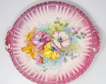 May include: A vintage pink and white porcelain serving plate with a floral design featuring pansies. The plate has a scalloped edge and two handles.