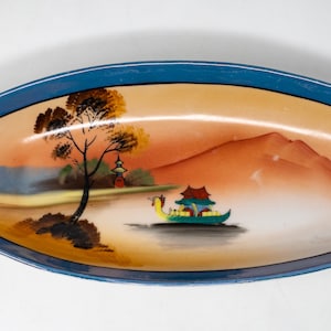 May include: An oval serving dish with blue trim and a painted scene of a mountain range, a pagoda, a tree, and a boat on a lake. The dish has two handles.