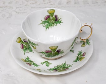 May include: A white teacup and saucer set with a floral pattern of green thistles. The cup has a gold rim and a gold handle.