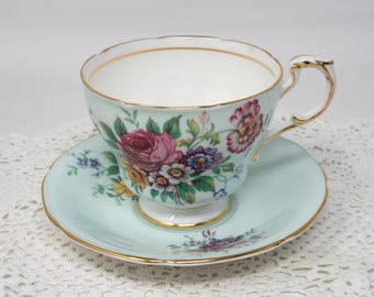 May include: A delicate teacup and saucer set with a floral pattern. The cup and saucer are a light blue color with a gold trim. The floral pattern features pink, yellow, and purple flowers.